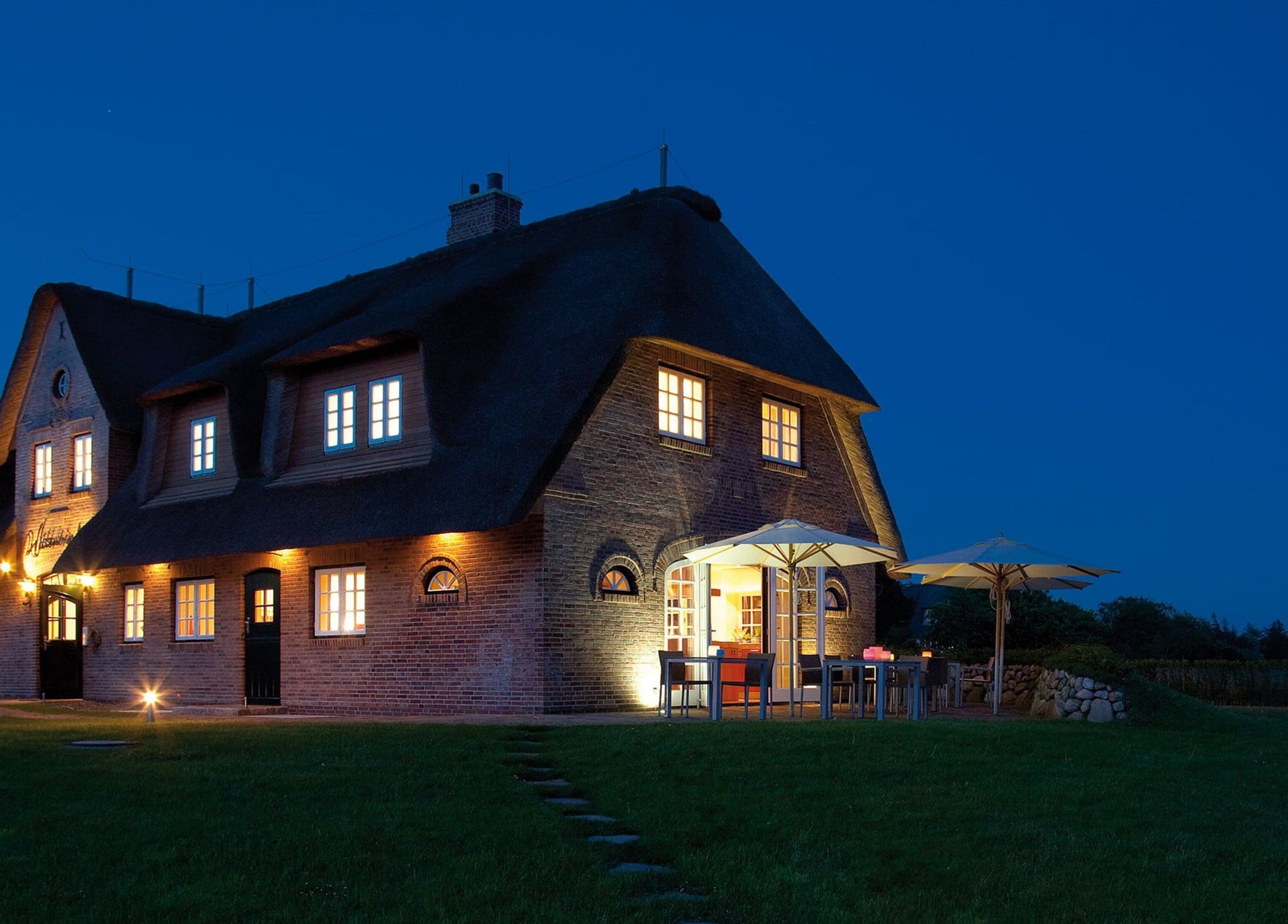 Reetgedecktes Backsteinhaus Lüüvhoog am Abend, beleuchtete Fenster und Terrasse mit Sonnenschirmen
