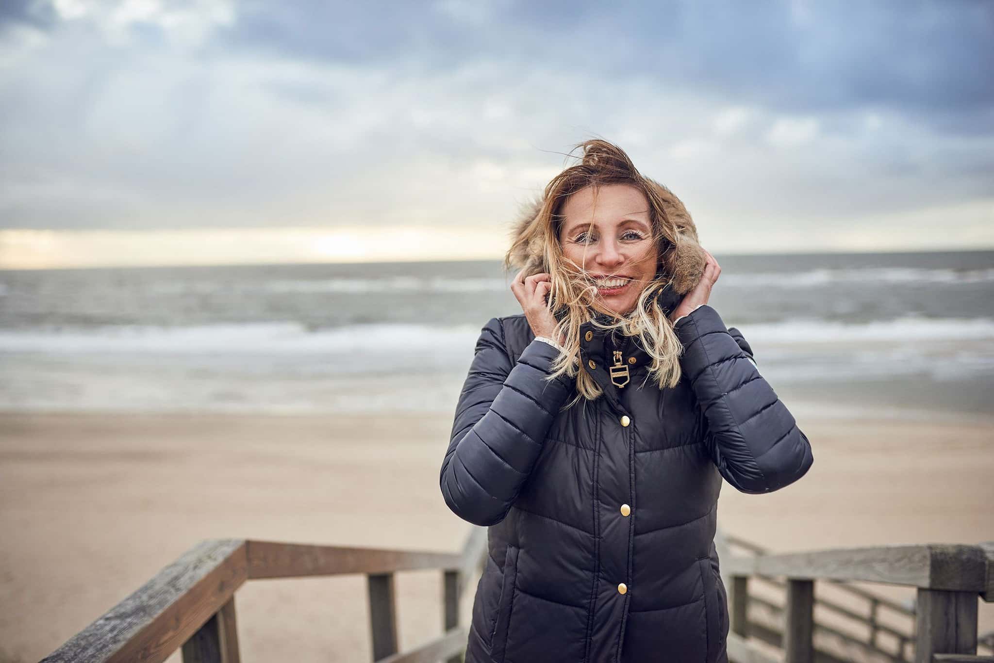 Frau in Winterjacke am windigen Strand, Holzgeländer, Wellen und bewölkter Himmel dahinter