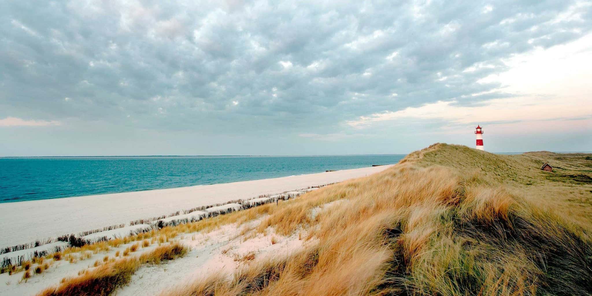 Dünenlandschaft mit Strandgras, hellem Sandstrand, Meer und rot-weißem Leuchtturm auf Sylt