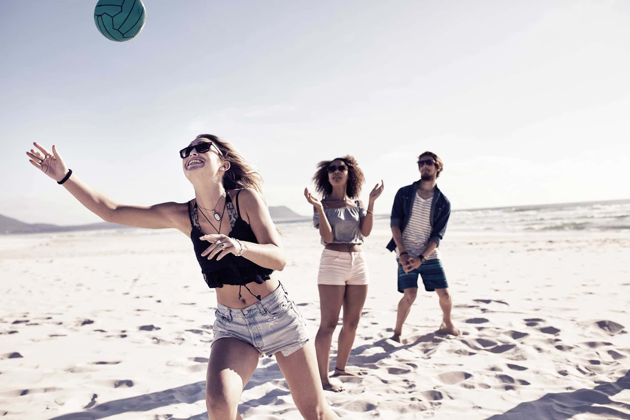 Freunde spielen am Strand Volleyball, eine junge Frau schlägt lachend den Ball