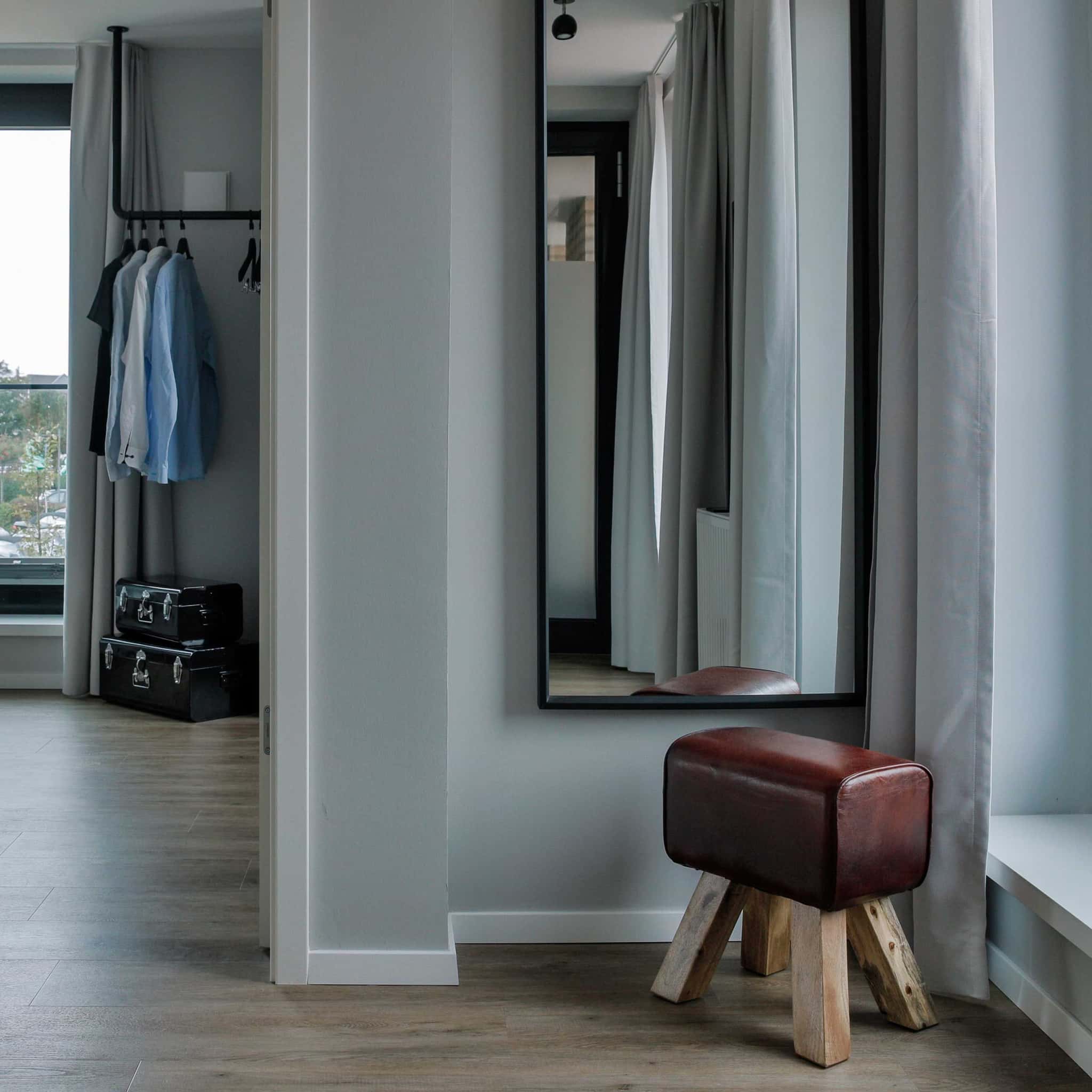 Garderobenstange mit Hemden, Koffer, großer Wandspiegel und Lederhocker im Appartement Bett & Bude