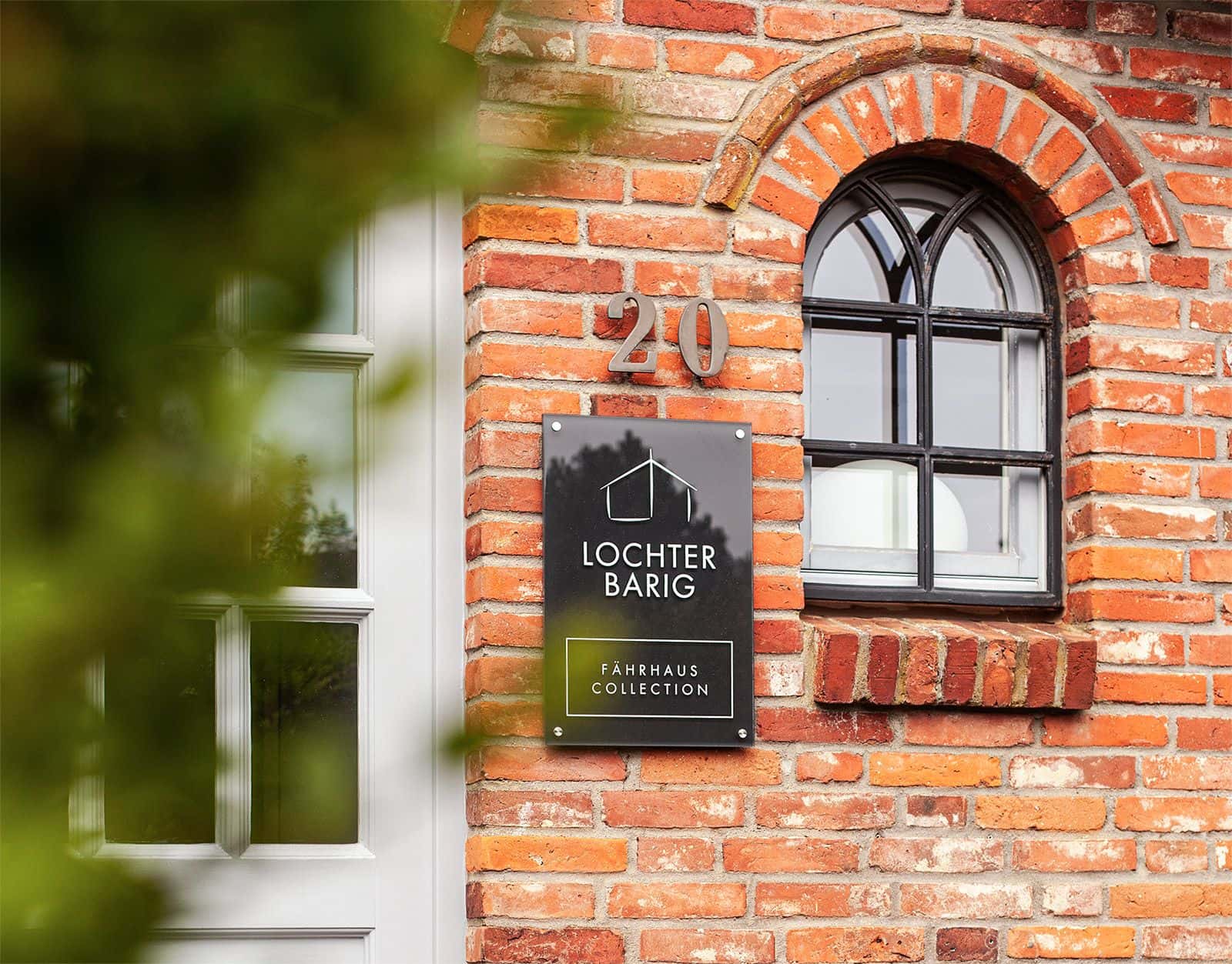 Backsteinfassade mit Rundbogenfenster, Hausnummer 20 und Namensschild Lochterbarig – Fährhaus Collection