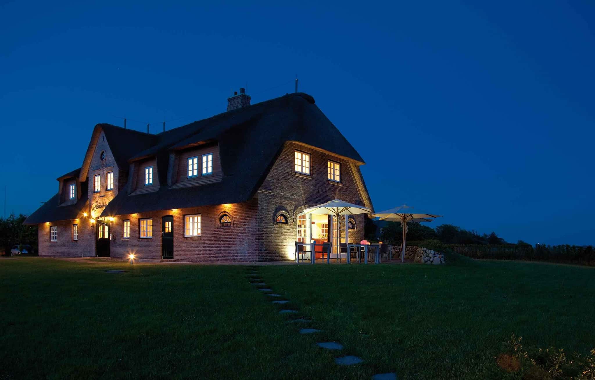 Reetgedecktes Backsteinhaus Lüüvhoog in der Dämmerung, beleuchtete Fenster, Terrasse und Rasen