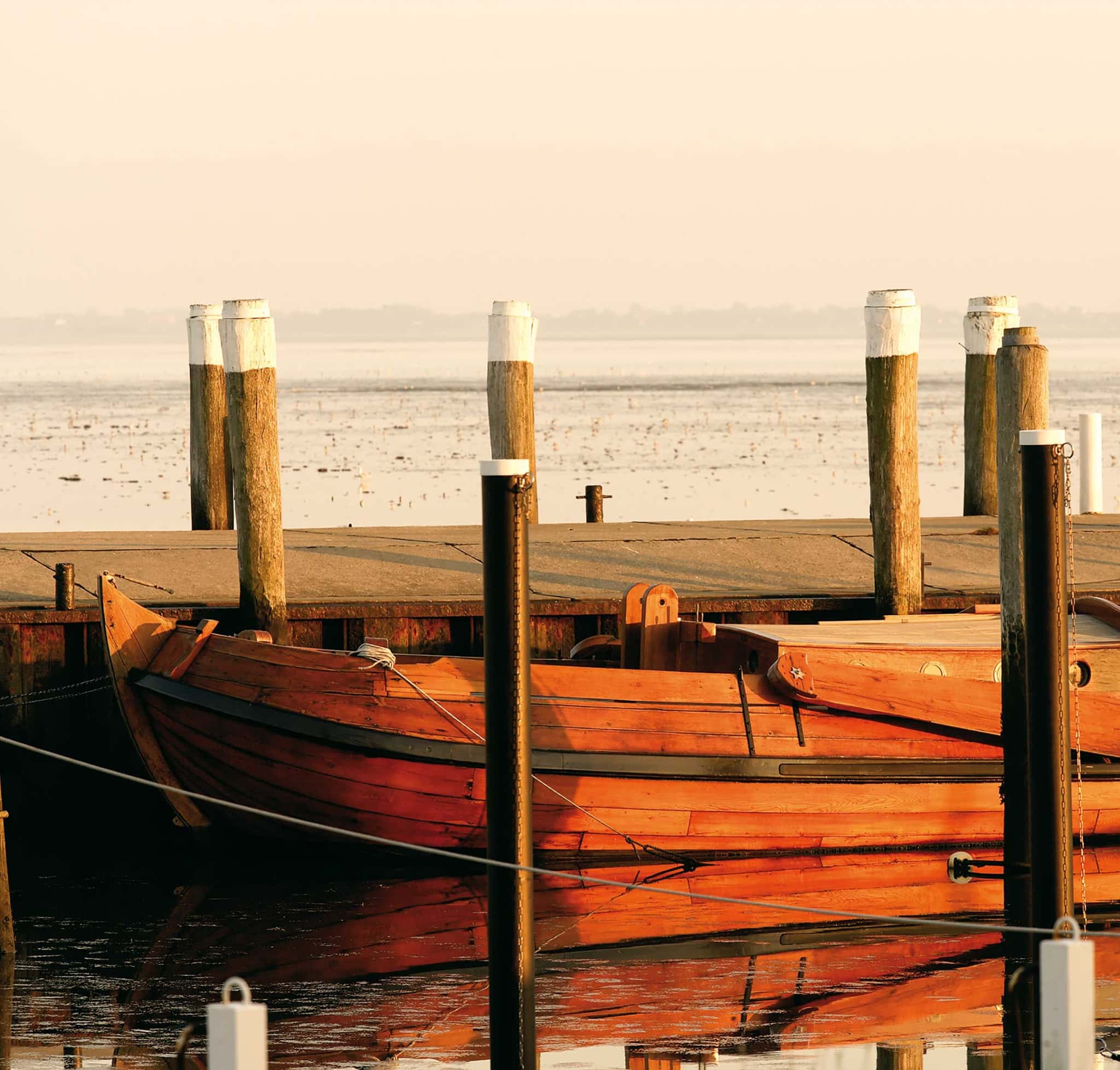 Holzboot am Anleger im Wattenmeer bei Morgenlicht, im Hintergrund Pfähle und weites Watt