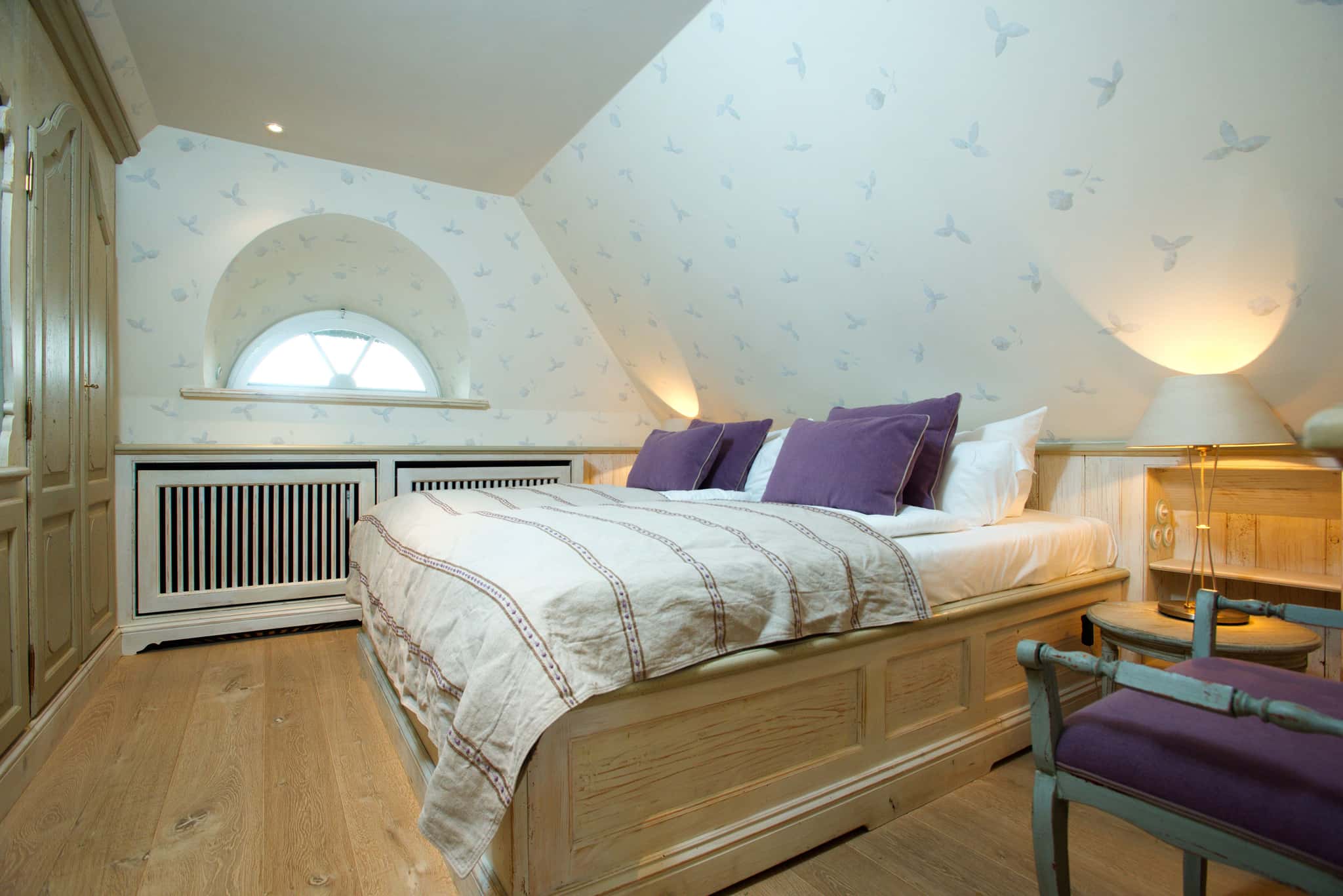Schlafzimmer mit Doppelbett, lila Kissen, Rundbogenfenster und gemusterter Tapete, Tipkenhoog